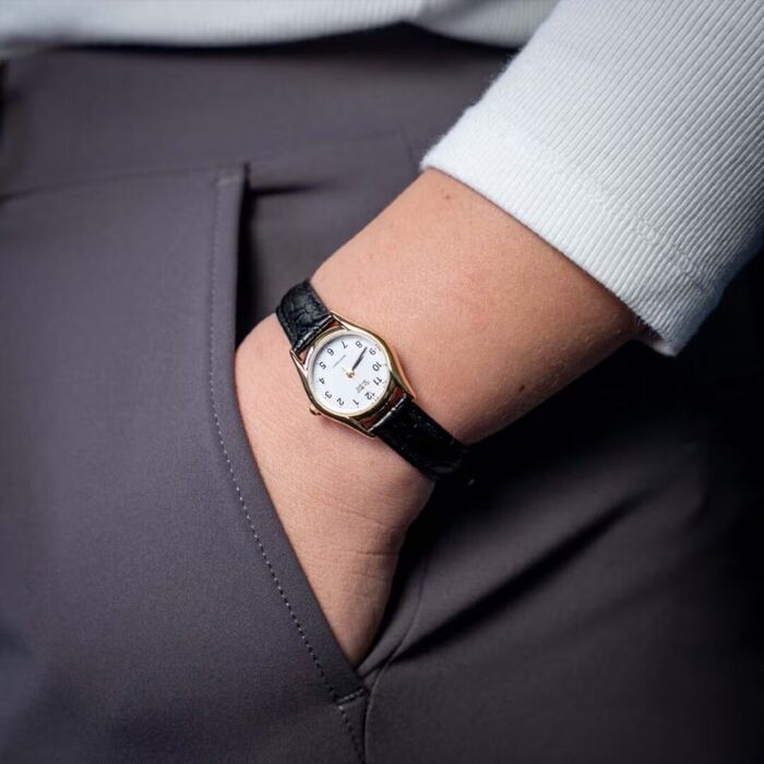 Pulso com relógio dourado de mostrador branco e bracelete preta, mão no bolso cinzento