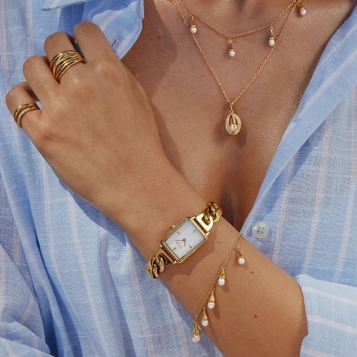 Modelo com joias e um relógio dourado One com uma caixa retangular e mostrador branco.