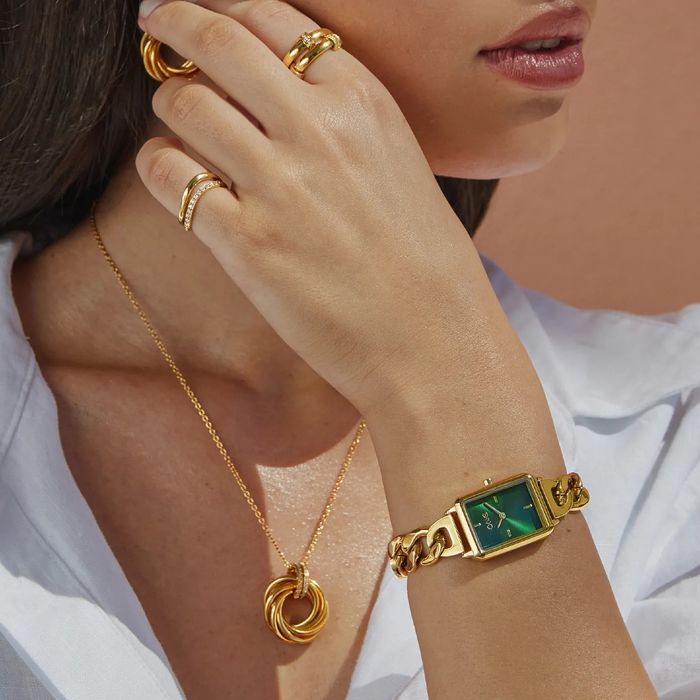 Modelo com colar, anéis e um relógio dourado One com uma caixa retangular e mostrador verde.