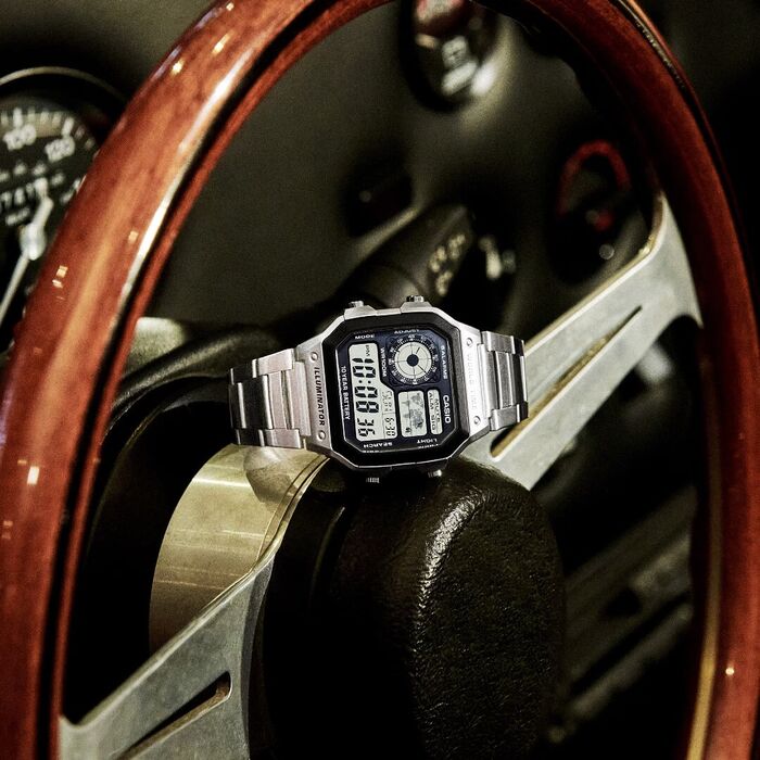 Relógio Casio em aço prateado com mostrador digital horário mundial, em cima de um volante do carro.