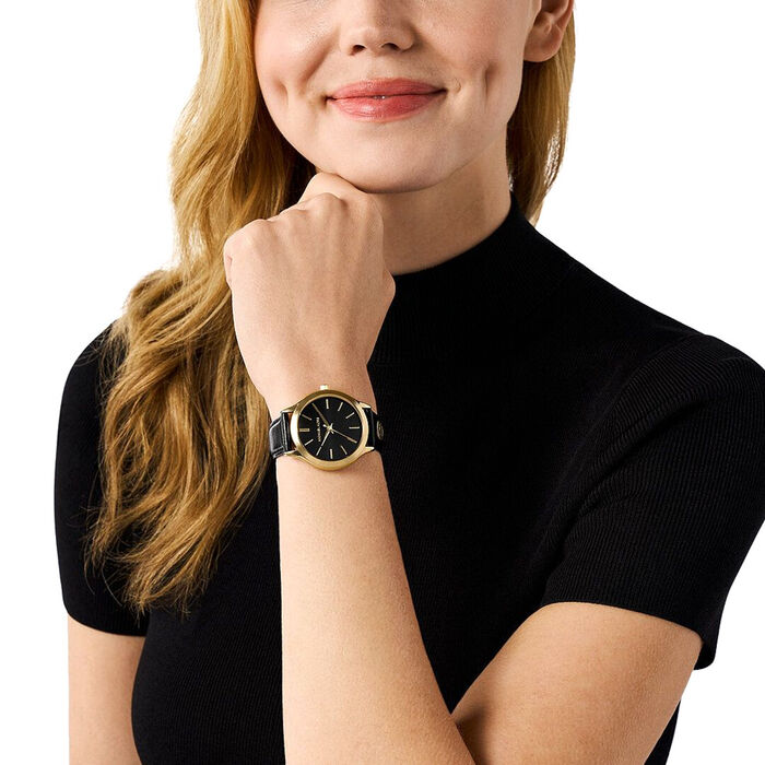 Relógio feminino da M.K com bracelete em couro preto, mostrador preto e caixa dourada, no pulso.