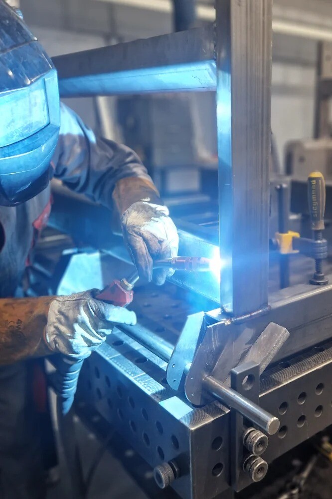 Welder performing manual welding on a steel part at a workbench, with the welding hall visible in the background, showcasing industrial metal fabrication.