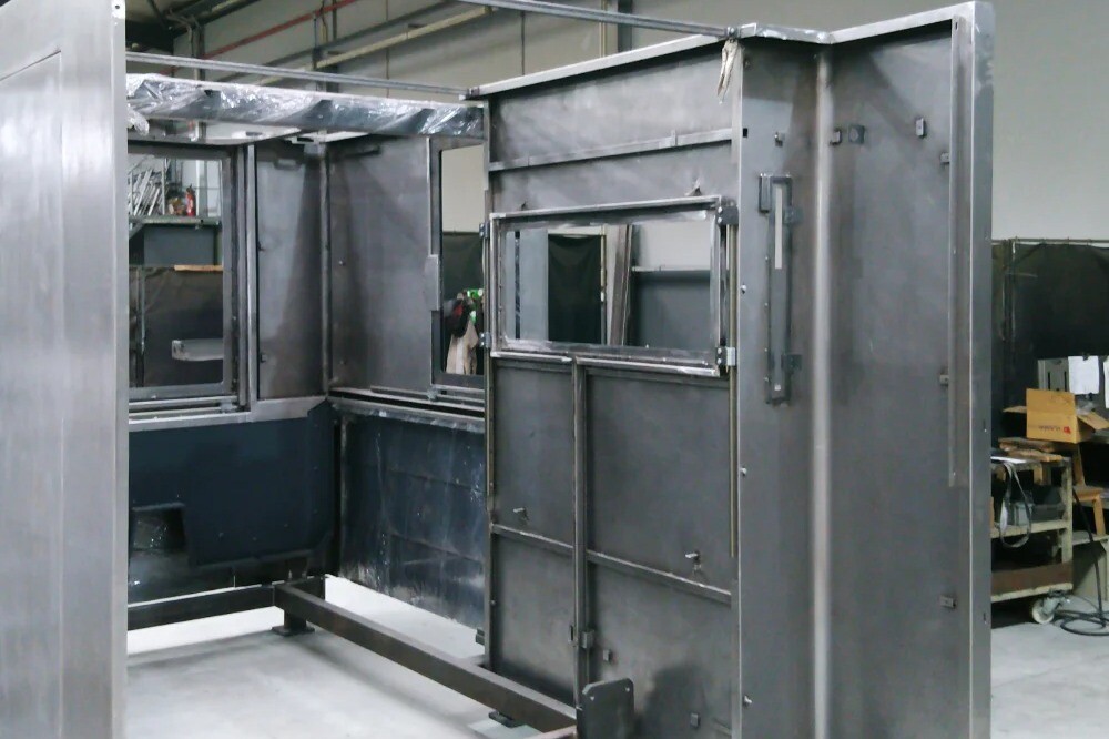 Large metal machine structure being welded inside an industrial warehouse with overhead crane, showing detailed fabrication process