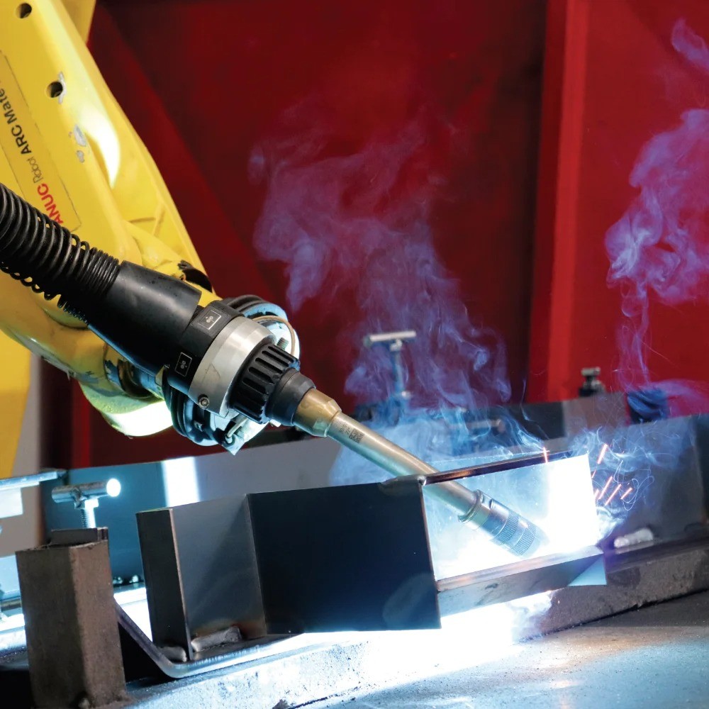 FANUC robotic welding cell performing MIG/TIG welding on a carbon steel structure at V Laser On in Porto Portugal, with welding sparks and smoke rising, showcasing automated high-precision industrial welding