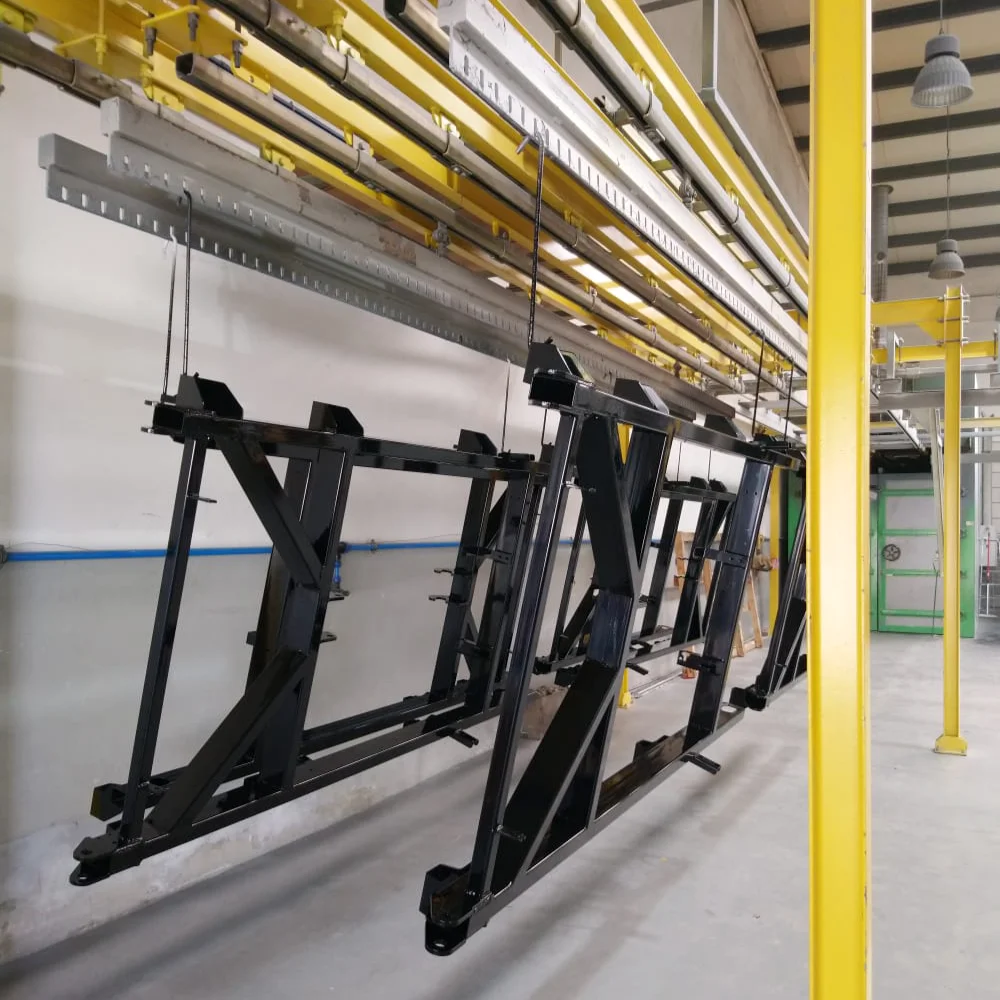 Black metal structures freshly painted, hanging on overhead rails and ready to enter the curing oven at V Laser On in Porto Portugal, showcasing industrial electrostatic powder coating workflow and oven polymerization process