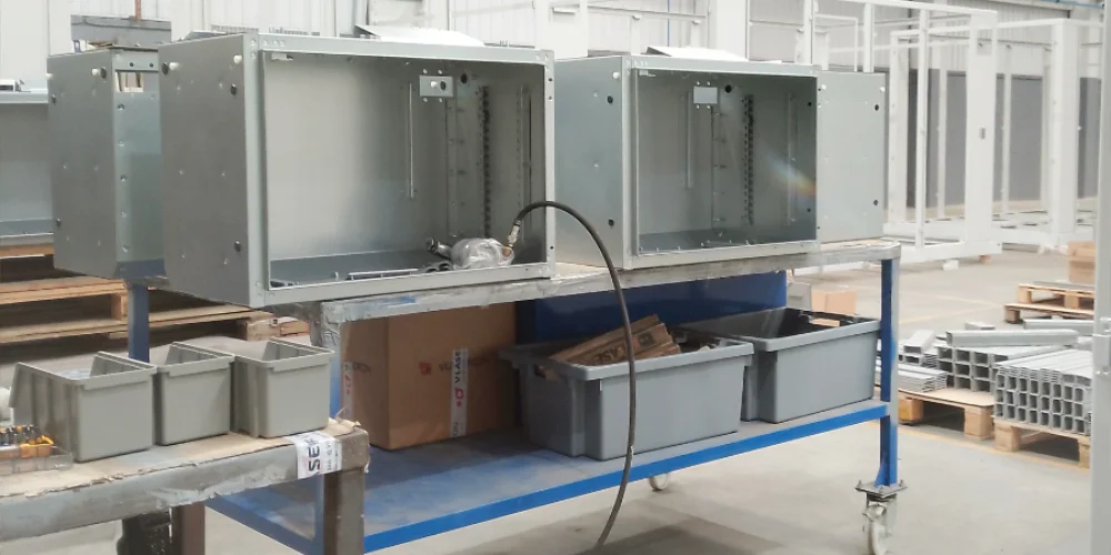 Electrical control boxes being assembled on a workbench at V Laser On in Porto Portugal, with welded metal structures visible in the background along the final product assembly line