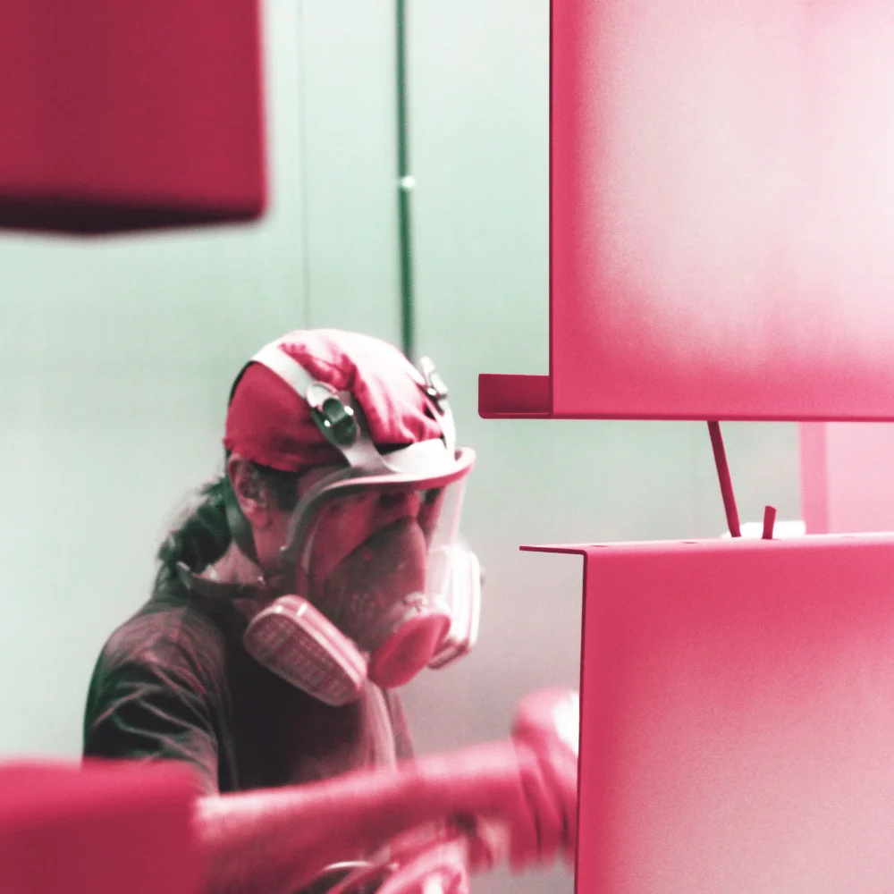 Red epoxy powder coating adhering to metal parts in the foreground, with a worker visible in the background at V Laser On workshop in Porto Portugal, demonstrating high-quality industrial coating process